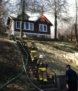 Firefighters climb stairs while pulling a water hose to a house fire on a small hill