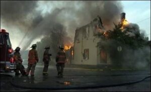 Three firefighters talk outside of a house fire while smoke rises from the flames and water can be seen spraying onto the burning structure