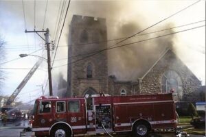 A fire engine outside of a church with a stone facade and bell tower while smoke surrounds the building