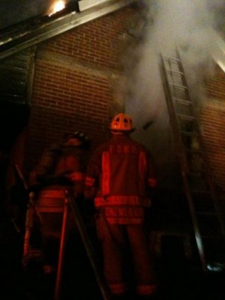 Firefighters at a fire at a brick building, with a ladder on the side of the building and smoking billowing out a hole