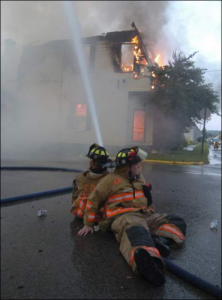 Two firefighters sit on the ground back-to-back to control a water hose while trying to combat a house fire in the background