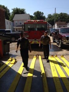 Two firefighters with a fire truck in the background and several hoses laid out for testing