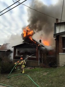 Three firefighters spray water onto a brick house with flames and smoke rising from the roof