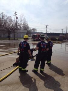 Three firefighters gather standing while two others are on the ground tending to a fire hose full of water during a training session